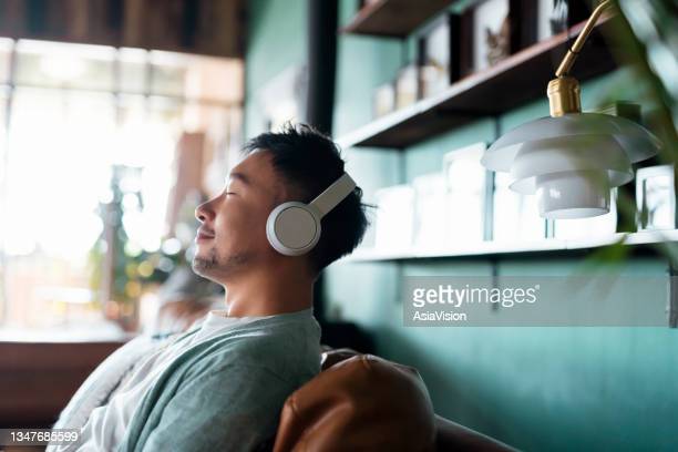Young Asian man with eyes closed, enjoying music over headphones while relaxing on the sofa at home