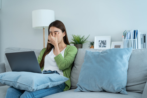 Exhausted Asian business woman hurt eye while using laptop computer. Young girl office worker sit on sofa feel visual fatigue and eye strain tired from overwork and massage dry irritable eye at home.