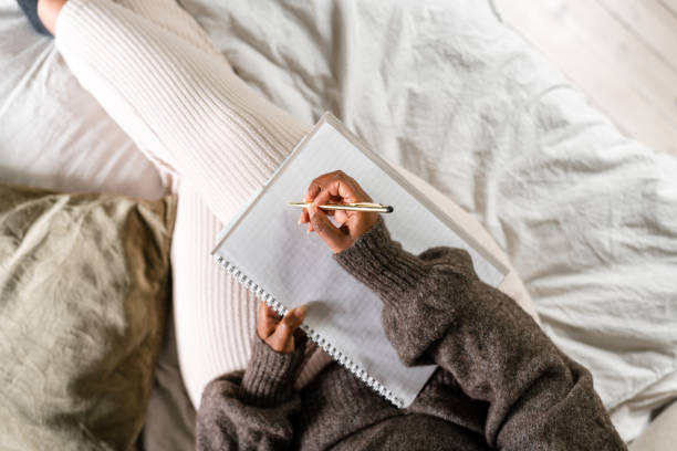 A woman is sitting on her bed, using a note pad to write. She is listening to something on her headphones.