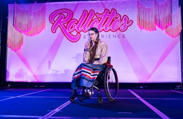 Cassidy is a lightskinned femme person sitting in their wheelchair. They are wearing brown moccasins with black and white fur on the ankles. They are wearing a grey ribbon skirt with black, white, and red ribbons. They are wearing a light tan cropper shirt. Their hair is on braids with brown mink fur tied to the ends. They are holding a microphone and are speaking into it. The background is pink with the words ROLLETTES EXPERIENCE in a darker pink.