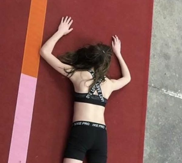 Image Description: a photo of Cassidy when they were nine years old, face down. They are fair skinned wearing a black sports bra and black sports shorts. They have long, brown, hair in a high ponytail. They are laying on a red mat that is next to grey concrete. To the left is a strip of red, pink, and orange.